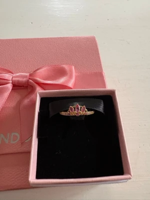 Guldfärgad ring med rosa stenar - Söt guldfärgad ring med rosa stenar i kronformad design. Ringen är original från Pandora och har inte blivit använd.