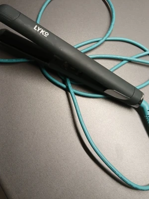 Svart plattång från Lyko - Säljer en svart plattång från Lyko med turkos sladd. Plattången har en stilren design och är enkel att använda för att styla håret. Perfekt för att skapa släta och glansiga frisyrer. Materialet är plast med metalliska detaljer.
