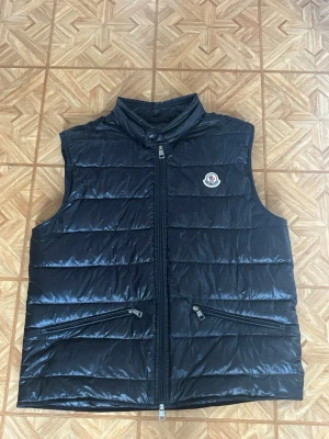 Moncler Väst - Snygg svart dunväst från Moncler med hög krage och två dragkedjefickor framtill. Västen har quiltad design och ett Moncler-märke på bröstet. Perfekt för lager-på-lager och håller dig varm under kyliga dagar. Varan är 100% äkta utan defekter, säljer pga av den är för liten🤩