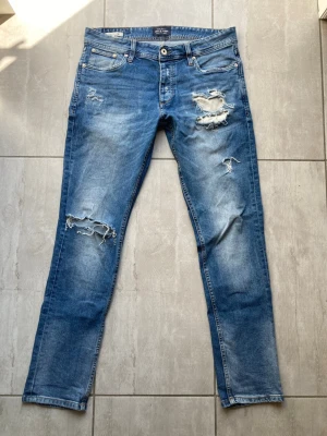 Jack n Jones jeans - Säljer ett par blå jeans med slitningar och hål på både lår och knä. Jeansen har fem fickor, knappgylf och raka ben. Perfekta för en avslappnad och trendig look. Materialet är klassisk denim i bomull. Väldigt eftertraktade på min TikTok strl 31