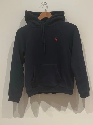 Ralph lauren - Snygg mörkblå hoodie från Polo Ralph Lauren med klassisk röd broderad logga på bröstet. Hoodien har huva med dragsko, stor magficka och ribbade muddar vid ärmar och nederkant. Perfekt för en avslappnad och stilren look. Storleken är ganska liten och passar barn runt 140 cm.