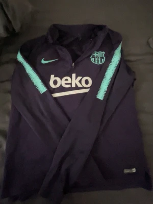 Barcelona träningströja Nike lila - Snygg lila långärmad träningströja från Nike med FC Barcelona-logga och turkosa detaljer på ärmarna. Halv dragkedja framtill och tryckt sponsorlogga 'beko' på bröstet. Perfekt för fotbollsträning eller chill.
