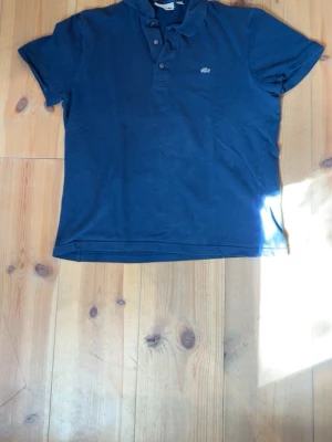Marinblå pikétröja från Lacoste - Säljer en marinblå pikétröja från Lacoste i slim fit-modell. Tröjan har korta ärmar, klassisk krage och knappar framtill. Ikoniska krokodilloggan på bröstet. Perfekt för en stilren och avslappnad look.