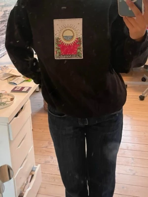 Svart hoodie med soltryck - Säljer en svart hoodie med ett färgglatt tryck på bröstet som föreställer en sol, himmel och blommor. Hoodien har en avslappnad passform och ribbade muddar vid ärmar och nederkant. Perfekt för dig som gillar unika prints och vill sticka ut. Passar både XS och S