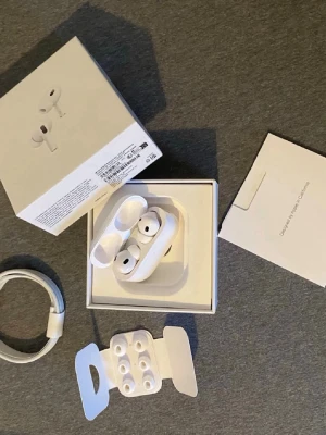 AirPods 2 Pro - Säljer ett par vita trådlösa in-ear hörlurar med laddningsetui, extra silikontoppar och laddningskabel. Hörlurarna har stilren design och levereras i originalförpackning med manualer och tillbehör. Perfekta för musik, poddar och samtal på språng.