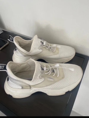 Vita  sneakers med textdetalj - Snygga vita sneakers med mesh-ovandel. Skorna har vita snören och en chunky sula. På hälen finns en svart textdetalj. Perfekta för en sportig och trendig look.