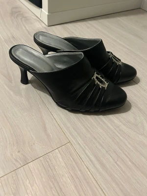 Svarta mules med dekorspänne - Säljer ett par svarta mules med smal klack och öppen häl. Skorna har dekorativa veck framtill och ett snyggt silverfärgat spänne med stenar på ovansidan. Materialet ser ut att vara konstläder och innersulan är silverfärgad. Perfekta för dig som gillar en chic och trendig look.