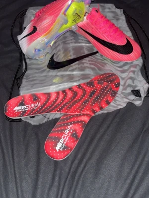 Nike Mercurial Superfly rosa elite fotbollsskor - Säljer ett par Nike Mercurial Superfly fotbollsskor i en grymt snygg rosa färg med svart swoosh. Skorna har en modern, strömlinjeformad design och genomskinlig yttersula med färgglada detaljer. Innersulorna är mönstrade i rött och svart. Perfekta för dig som vill sticka ut på planen.
