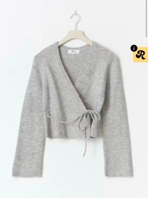 Grå omlottkofta med knytband - Mysig grå omlottkofta i mjukt material med långa ärmar och knytband i midjan. Perfekt för lager på lager och enkel att styla till både jeans och kjol. Koftan har en croppad passform och v-ringad hals.