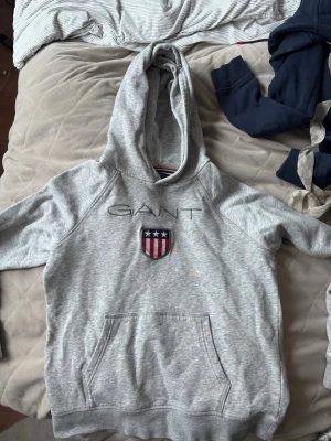 Grå hoodie från GANT med logga - Snygg grå hoodie från GANT med broderad logga och sköld med stjärnor och ränder på bröstet. Klassisk känguruficka framtill och huva med dragsko. Mjuk bomullsblandning och avslappnad passform. Perfekt för en casual look.