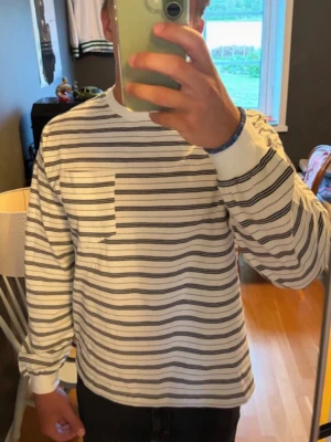 Vit långärmad randig tröja - Snygg vit långärmad tröja med svarta och gråa horisontella ränder. Tröjan har rund halsringning, ribbade muddar och en liten bröstficka på vänster sida. Perfekt för en avslappnad och stilren look.