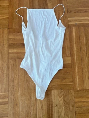 Vit ribbad body med smala axelband - Snygg vit body med ribbad struktur och smala axelband. Modellen har raka kanter upptill och högt skurna ben. Perfekt att styla till jeans eller kjol för en trendig look.