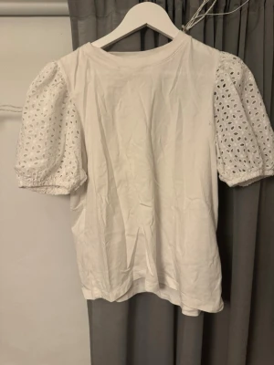 Vit topp med puffärm och broderade detaljer - Säljer en vit topp med puffiga, korta ärmar i broderad spets och elastisk kant. Kroppen är i slät bomull och toppen har en rund halsringning. .