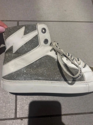 Glittriga  sneakers från Zadig&Voltaire - Snygga höga sneakers i vitt och silver med glittrande tyg och en cool blixtdetalj på sidan. Skorna har vita snören med text och dragkedja på insidan för enkel påtagning. Perfekta för dig som vill sticka ut med din stil.