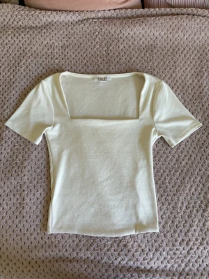 Vit topp med fyrkantig ringning - Snygg vit topp med korta ärmar och fyrkantig ringning. Toppen har en slim passform och är tillverkad i ett mjukt, stretchigt material som känns skönt mot huden. Perfekt att styla med jeans eller kjol för en clean look.