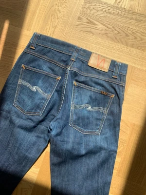 Nudie Jeans Slim Jim - Snygga Jeans i Nudie i modellen Slim Jim. Jeansen har en slim bootcut passform och är I storlek 29. Den korta loggan på bakfickan indikerar på en äldre modell från Nudie som ej säljs längre. Mått, Midja: 40 Ytterben: 102 Innerben: 79 Benöppning: 19