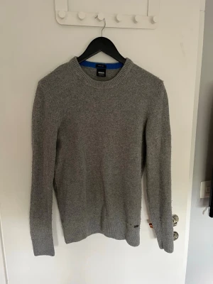 Hugo Boss stickad tröja - Säljer en stilren grå stickad tröja från Hugo Boss. Tröjan har en klassisk rund halsringning och långa ärmar. Den har en subtil logga vid nederkanten. Storlek M men känns som S