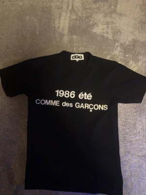 Svart Comme des Garçons t-shirt - Svart t-shirt från Comme des Garçons med vit text '1986 été' och logga framtill. Den är storlek M men passar som S, säljer den eftersom att den är passar för liten på mig. 