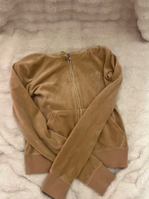Beige velourhoodie från Juicy Couture - Säljer en beige velourhoodie från Juicy Couture med dragkedja framtill och känguruficka. Klassisk broderad logga på bröstet och ribbade muddar vid ärmslut och nederkant. Perfekt för en avslappnad och trendig look.