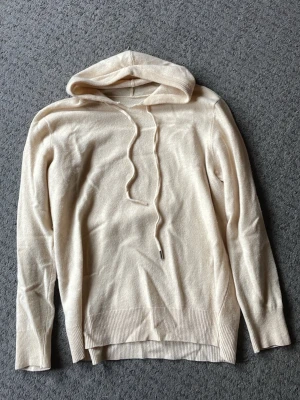 Merinoull hoodie - Snygg beige stickad hoodie med huva och dragsko. Tröjan har långa ärmar och ribbade muddar vid ärmslut och nederkant. Perfekt för en chill och avslappnad stil. Materialet känns mjukt och skönt mot huden. En av snörerna är trasigt längst ner och sömmen har lossnat lite vid halsen men annars bra skick i övrigt