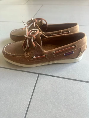 Bruna seglarskor från Sebago dam - Klassiska bruna seglarskor från Sebago i mocka med vita gummisulor och råa lädersnören. Skorna har dekorativa sömmar och metallöljetter samt en liten Sebago-logga på sidan. Perfekta för en avslappnad och stilren look. Helt nya!