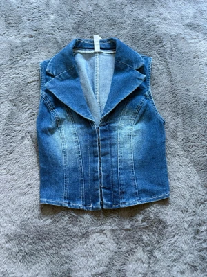 Ärmlös jeansväst med bred krage - Säljer en snygg ärmlös jeansväst i klassisk blå denim med bred krage och markerade sömmar framtill. Västen har figurnära passform och stängs framtill. Perfekt att styla över en t-shirt eller skjorta för en cool look.