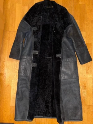 Hollies äkta fårskinnskappa – svart vintage – storlek 38 - Äkta fårskinnskappa från Hollies i klassisk lång modell, storlek 38. Tillverkad i äkta fårskinn med varm och mjuk päls på insidan. Kraftig och gedigen kappa med stora fickor framtill och en tidlös vintage-design.  Färgen är svart/mörkgrå med naturlig patina som ger kappan en fin och unik karaktär. Kappan är i mycket bra skick.  Storlek: 38 Material: Äkta fårskinn Färg: Svart/mörkgrå Skick: Mycket bra skick 