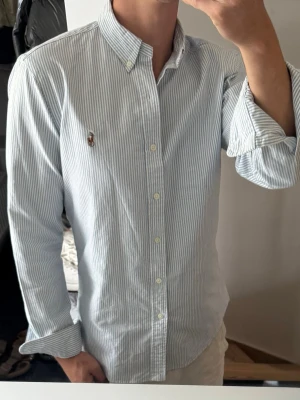 Blårandig skjorta med knappar - Snygg skjorta med tunna blå och vita ränder, klassisk button-down krage och knappar framtill. Skjortan har lång ärm och en liten broderad detalj på bröstet. Perfekt för en clean och stilren look. Storlek m men sitter som S