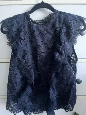 Mörkblå spetsblus med volangärm - Snygg mörkblå blus i spets med blommigt mönster och volangdetaljer vid ärmarna. Blusen är ärmlös med rund halsringning och har en feminin, romantisk look. Perfekt att styla till jeans eller kjol för en trendig outfit.