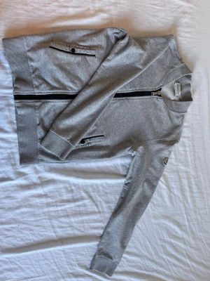 Grå ziptröja med ribbade muddar - Snygg grå ziptröja i sweatshirtmaterial med svarta dragkedjor och två fickor framtill. Tröjan har ribbade muddar vid ärmslut och nederkant samt en patch på ena ärmen. Klassisk och enkel design utan huva.