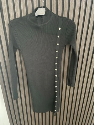 Svart ribbad stickad klänning - Snygg svart ribbad stickad klänning med lång ärm och hög krage. Klänningen har dekorativa knappar längs ena sidan framtill som ger en cool detalj. Perfekt till höst och vinter för en stilren look.