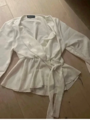 Vit omlottblus med knytband - Snygg vit omlottblus med knytband i midjan och långa ärmar. Blusen har en avslappnad passform och volangkant nedtill. Perfekt att styla med jeans eller kjol för en trendig look.