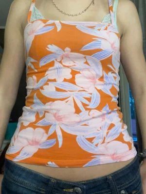 Orange blommig linnetopp - Säljer en färgglad orange linnetopp med blommigt mönster i ljusrosa och lila toner. Toppen är ärmlös med smala axelband och har en figurnära passform. Perfekt för varma dagar och somriga outfits! Storlek är ca XS / S