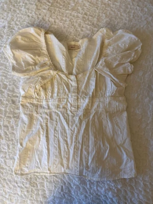 Vit blus med puffärm från Nelly - Söt vit blus från Nelly med puffiga korta ärmar och v-ringning. Blusen har knappar framtill och smock i ryggen för en skön passform. Tillverkad i ett lätt, strukturerat tyg som ger en luftig känsla. Perfekt till jeans eller kjol.