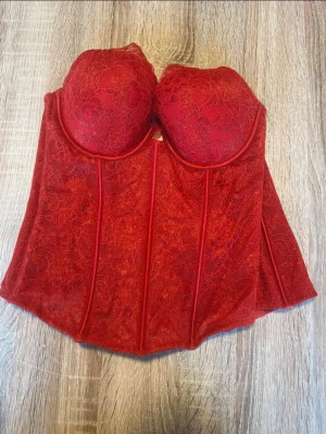 Röd spetskorsett med bygel - Snygg röd korsett med vadderade kupor och bygel för extra stöd. Hela korsetten är täckt av blommig spets och har markerade sömmar för en klassisk siluett. Materialet är genombrutet och ger en lyxig känsla.