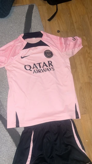 PSG rosa och svart matchställ Nike - Säljer ett Paris Saint-Germain matchställ från Nike i rosa och svart. Tröjan är kortärmad med PSG-logga och Qatar Airways-tryck framtill. Shortsen är svarta med rosa detaljer och PSG-märke. Perfekt för fotbollsträning eller match!