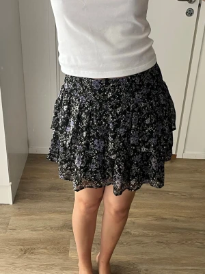Svart blommig volangkjol - Kort svart kjol med blommigt mönster i lila och vitt. Kjolen har flera volanger och resår i midjan för en bekväm passform. Köpt på barnavdelningen men passar mig som vanligtvis ha storlek S
