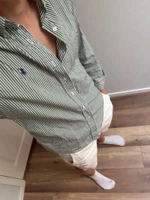 Grönvit randig skjorta från Polo Ralph Lauren - Snygg grön och vit randig skjorta från Polo Ralph Lauren med klassisk button-down krage och blå logga broderad på bröstet. Skjortan har långa ärmar och knäppning framtill. Perfekt till jeans eller linneshorts för en clean look.