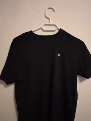 Svart t-shirt från GANT - Klassisk svart t-shirt från GANT med liten broderad logga på bröstet. T-shirten har rund hals och korta ärmar, perfekt till vardagslooken. Tillverkad i mjuk bomull för skön känsla hela dagen.