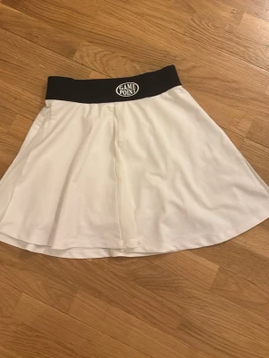 Vit kortkjol från Gina Tricot - Säljer en vit kortkjol från Gina Tricot med svart bred resår i midjan och trycket 'Game Point' framtill. Kjolen har en sportig look och är perfekt för tennis eller andra aktiviteter. Materialet känns lätt och bekvämt.