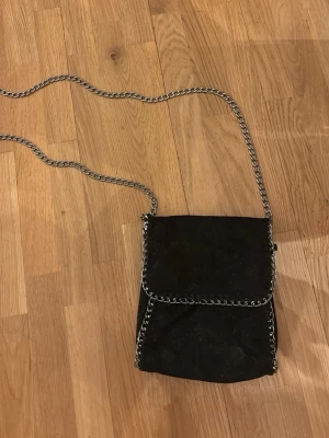 Svart axelväska med kedjedetaljer - Snygg svart axelväska med silverfärgad kedja som axelrem och runt kanterna. Väskan har lock med magnetknäppning och ett innerfack med dragkedja. Materialet ser ut som mockaimitation och ger en cool vibe till outfiten.