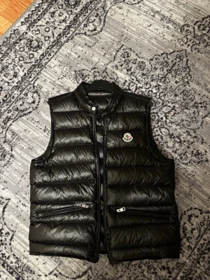 Moncler gui väst - Snygg svart Moncler gui väst med glansig finish och klassisk logotyp på bröstet. Västen har ståkrage, två dragkedjeförsedda fickor och quiltad design. Perfekt för lager-på-lager och streetstyle. Priset är ej hugget i sten så var inte rädda att ställa frågor 😁