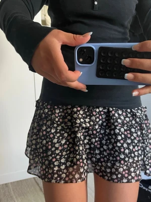 Svart blommig volangkjol från Sofie schnoor  - Kort svart kjol med volanger och små vita och rosa blommor. Kjolen har ett tunt, lätt material och sitter högt i midjan. Perfekt för dig som gillar en söt och trendig stil.