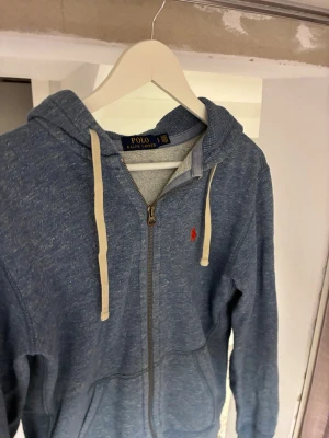 Blå zip hoodie från Polo Ralph Lauren - Blåmelerad hoodie med dragkedja från Polo Ralph Lauren. Klassisk huva med vita snören och röd broderad logga på bröstet. Mjuk insida och kängurufickor framtill. Perfekt för en avslappnad och stilren look.