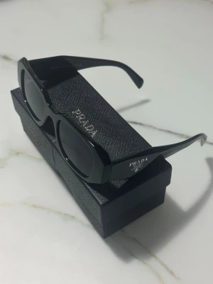 Prada solglasögon - Snygga svarta solglasögon från Prada med rektangulära bågar och diskret logga på sidan. Glasögonen har en stilren och modern design som passar perfekt till alla outfits. Kommer med originalfodral.