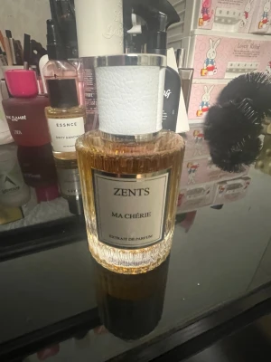 Zents Ma Chérie Eau de Parfum - Zents Ma Chérie Eau de Parfum i en elegant, genomskinlig glasflaska med räfflad botten och vit texturerad kork med silverdetaljer. Doften är exklusiv och flaskan har en lyxig känsla som passar perfekt på sminkbordet.