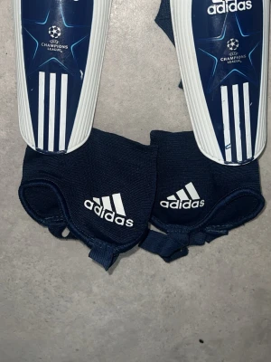Adidas benskydd Champions League - Säljer ett par mörkblå och vita Adidas benskydd med Champions League-logga. Klassisk design med tre vita ränder och stjärndetaljer. Tillhörande elastiska fotremmar med Adidas-logga. Perfekta för fotbollsträning eller match.