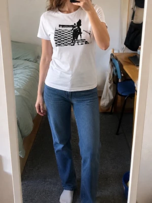 Vit t-shirt med svart tryck - Vit t-shirt med ett svart grafiskt tryck av en byggnad och en ryttare på häst. Klassisk rund hals och korta ärmar. Passformen är normal och materialet känns som mjuk bomull. Perfekt för dig som gillar unika motiv och enkel stil.