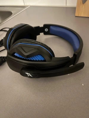 Svart och blått gamingheadset med mikrofon - Säljer ett gamingheadset i svart och blått med vadderad bygel och stora over-ear kåpor. Headsetet har inbyggd mikrofon och ansluts med kabel. Designen är robust med detaljer i plast och tyg för extra komfort. Perfekt för gaming eller videosamtal. Ska funka till alla konsoler.