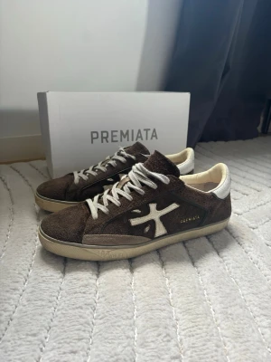 Bruna Premiata sneakers i mocka - Snygga bruna sneakers från Premiata i mjuk mocka med vita detaljer och logga på sidan. Perfekta för dig som gillar stilrena och trendiga sneakers. Använt fåtal gånger näst in till nya.